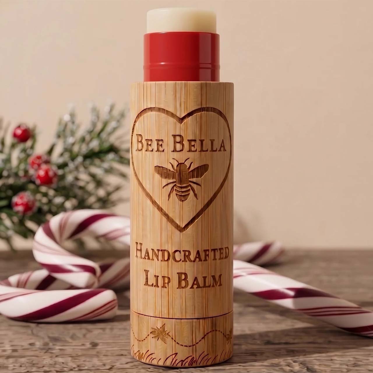 A lip balm stick in a bamboo-textured tube with a red cap, labeled 'BEE BELLA HANDCRAFTED LIP BALM,' standing on a wooden surface with candy canes and evergreen branches in the background.