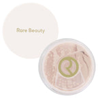 Rare Beauty loose setting powder in a clear round compact with the cream-colored lid placed behind it on a white background.