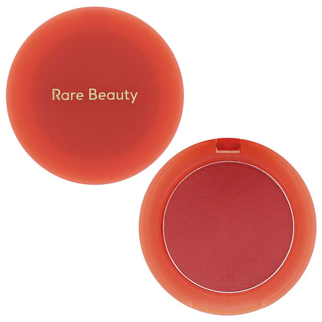 Rare Beauty round makeup compact shown open with the lid beside it on a white background. The compact is translucent coral-orange, and the pressed powder inside is a warm rosy red.