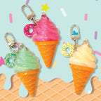 Three ice cream cone keychains with silver clasps, featuring pink, mint green, and pale yellow swirls in waffle cones with colorful sprinkle donut charms, set against a light blue background with pastel confetti and waffle graphics.