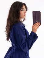 A woman with long dark brown hair, wearing a navy blue long-sleeve shirt, holds up a dark purple textured zip-around wallet with card slots and a gold snap button against a plain white background.